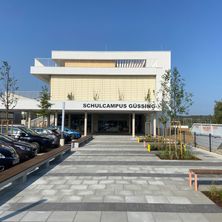 Das Schulcampus Gussing-Gebäude befindet sich in einer Stadt in Österreich, vor dem Gebäude sind Autos geparkt, und es gibt Bänke und Pflanzen im Vorgarten.