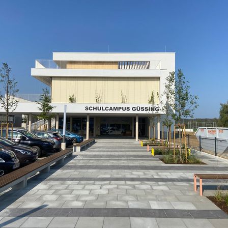 Das Schulcampus Gussing-Gebäude befindet sich in einer Stadt in Österreich, vor dem Gebäude sind Autos geparkt, und es gibt Bänke und Pflanzen im Vorgarten.