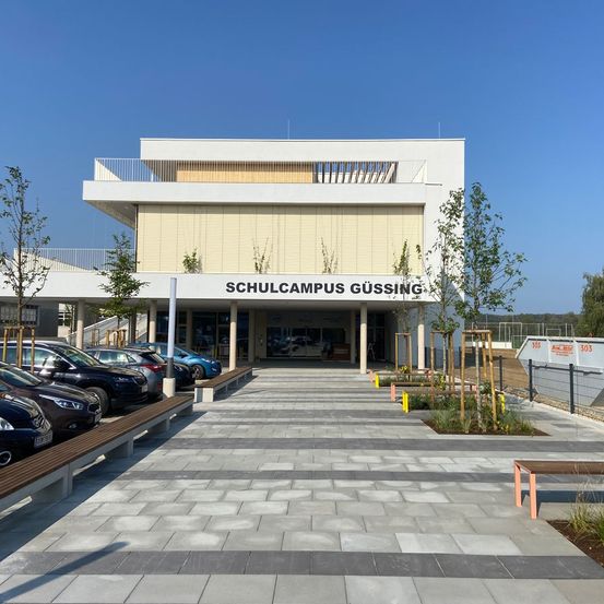 Das Schulcampus Gussing-Gebäude befindet sich in einer Stadt in Österreich, vor dem Gebäude sind Autos geparkt, und es gibt Bänke und Pflanzen im Vorgarten.
