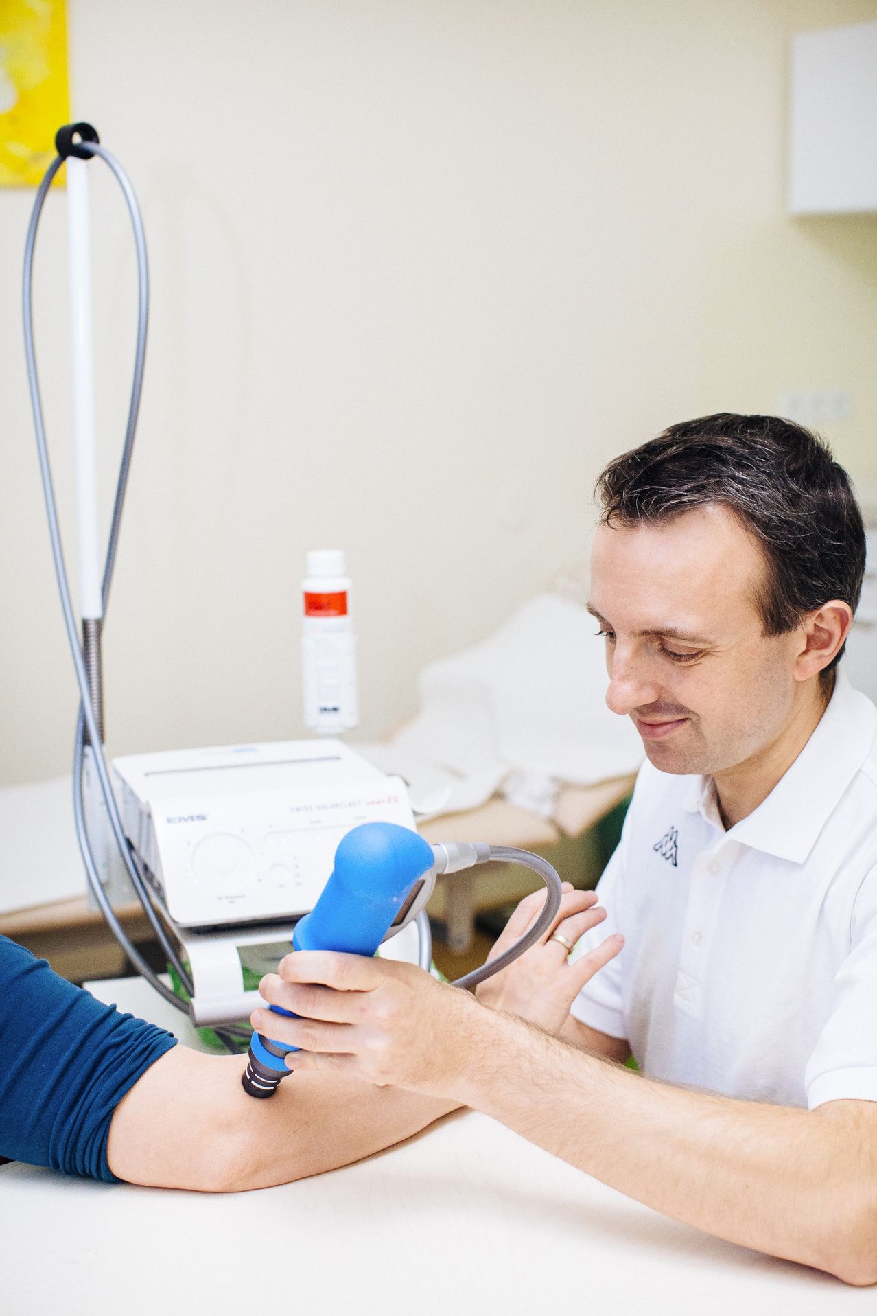 Ein männlicher Gesundheitsarbeiter in einem weißen Poloshirt verwendet ein blaues Ultraschallgerät am Arm eines Patienten. Das Gerät ist an eine Maschine mit elektronischem Display angeschlossen, und eine weiße Flasche ist darauf platziert.