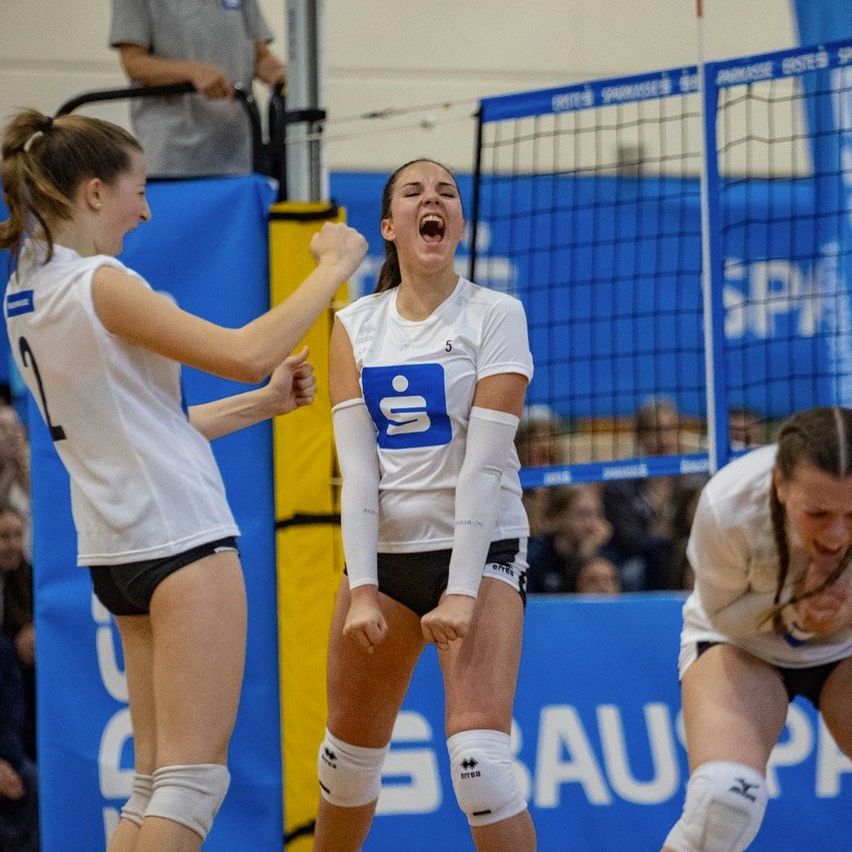 Drei Volleyballspielerinnen in weißen Trikots feiern einen erfolgreichen Spielzug. Eine Spielerin in der Mitte schreit mit offenem Mund. Die Spielerinnen befinden sich in einer Halle mit einem Netz und einer Menge im Hintergrund.