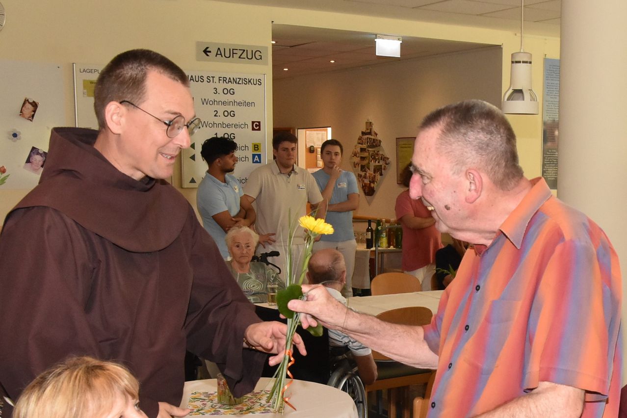 Ein Mann in einem braunen Gewand überreicht einem anderen Mann in einem gestreiften Hemd eine gelbe Blume. Im Hintergrund stehen und sitzen weitere Personen, darunter eine Frau im Rollstuhl.