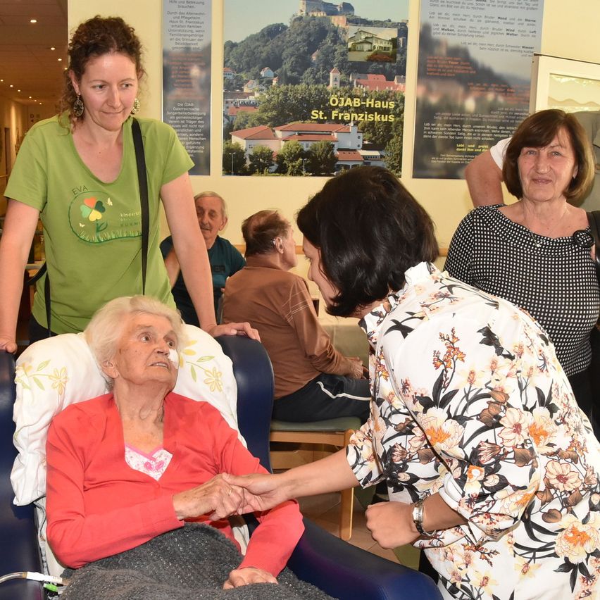 Eine ältere Frau sitzt in einem Stuhl, während eine Frau in einem Blumenhemd ihre Hand hält. Eine Frau in einem grünen Shirt steht hinter ihnen. Andere Menschen sitzen im Hintergrund.