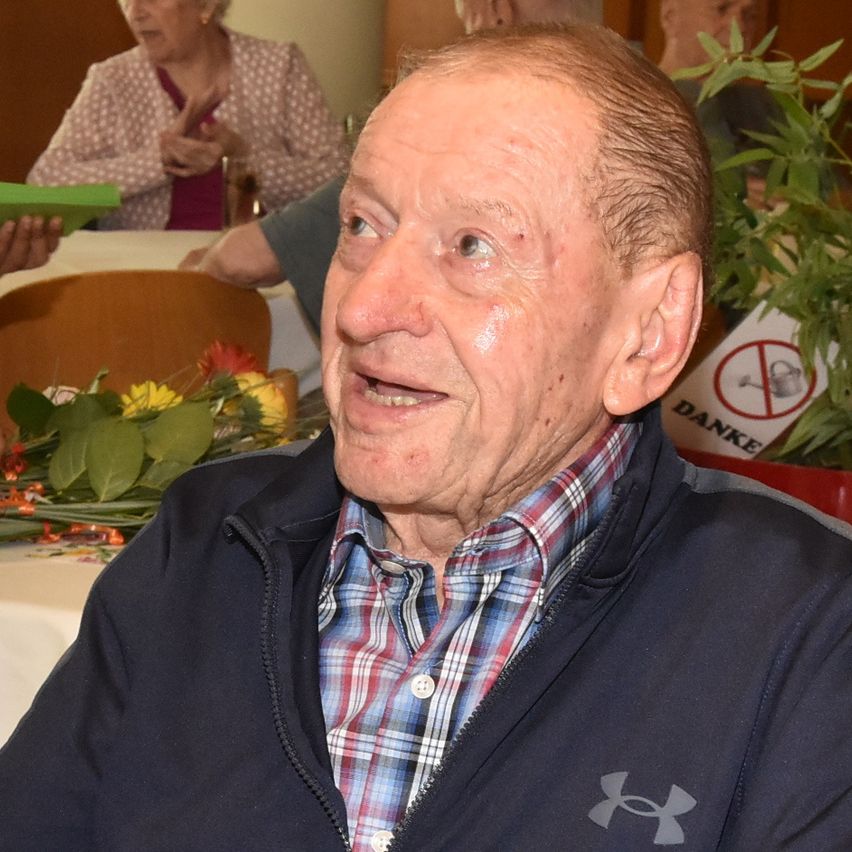 Ein älterer Mann lächelt und schaut nach vorne, trägt ein kariertes Hemd und eine dunkelblaue Jacke. Hinter ihm deutet eine Frau mit ihren Händen.