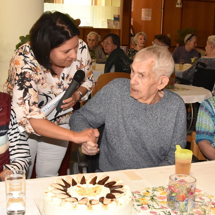 Ein älterer Mann sitzt an einem Tisch mit einem Kuchen, während eine Frau ihm ein Mikrofon reicht. Sie sind von anderen älteren Menschen in einem Raum umgeben.