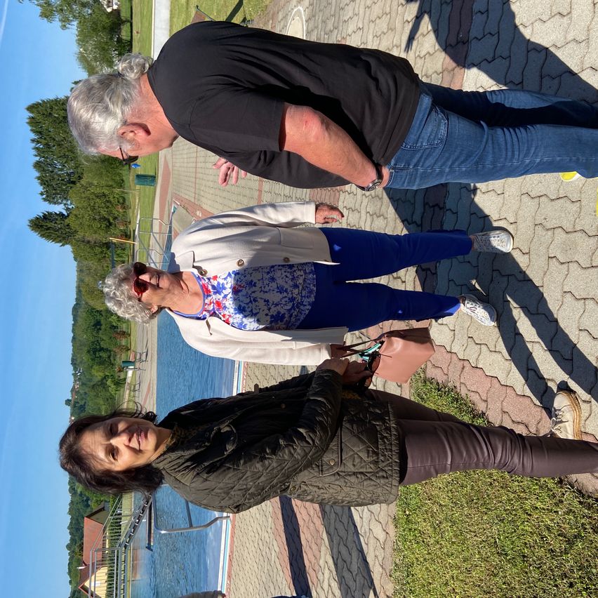 Drei Menschen stehen in der Nähe eines Gewässers. Ein älterer Mann schaut nach unten, während eine ältere Frau mit einem Blumenhemd lächelt. Eine weitere Frau in einer Jacke schaut zur Seite.