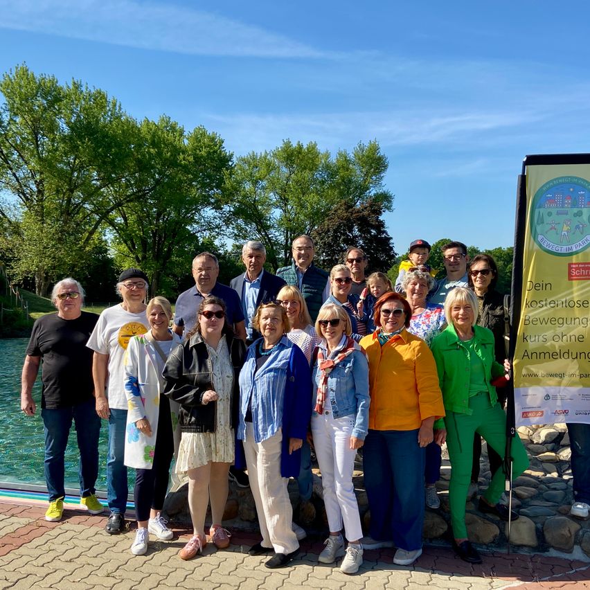 Eine Gruppe von Erwachsenen und Kindern steht vor einem See und lächelt für ein Foto. Der Hintergrund zeigt Bäume und einen klaren blauen Himmel.