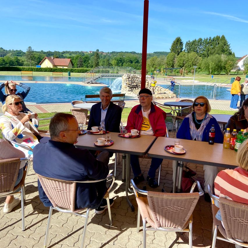 Eine Gruppe von Menschen sitzt draußen um Tische herum, in der Nähe eines großen Pools. Sie sind lässig gekleidet, einige tragen Brillen. Die Tische haben Tassen und Teller. Im Hintergrund befinden sich Bäume und Häuser.