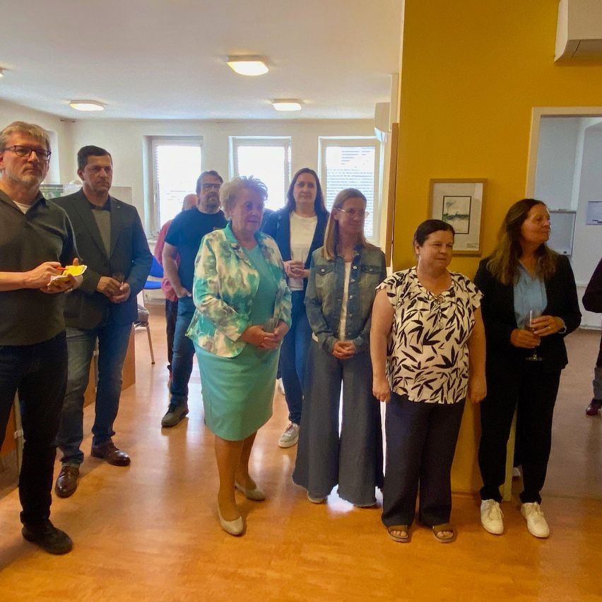 Eine Gruppe von Menschen steht in einem Raum mit Holzboden und gelben Wänden. Sie lächeln und scheinen sich in einer ungezwungenen Umgebung zu befinden.