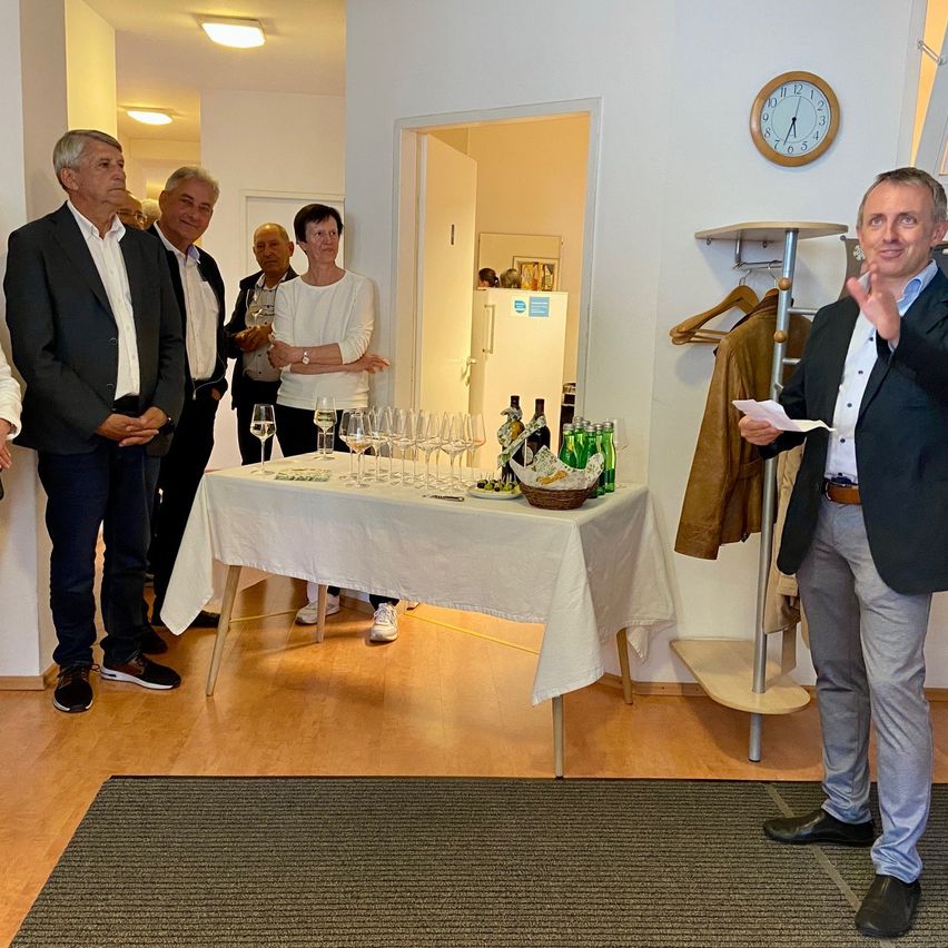 Eine Gruppe von Menschen in formeller Kleidung steht um einen Tisch mit Weingläsern und Flaschen. Ein Mann steht davor, hält ein Papier und spricht. Eine Uhr und ein Kleiderständer sind im Hintergrund zu sehen.