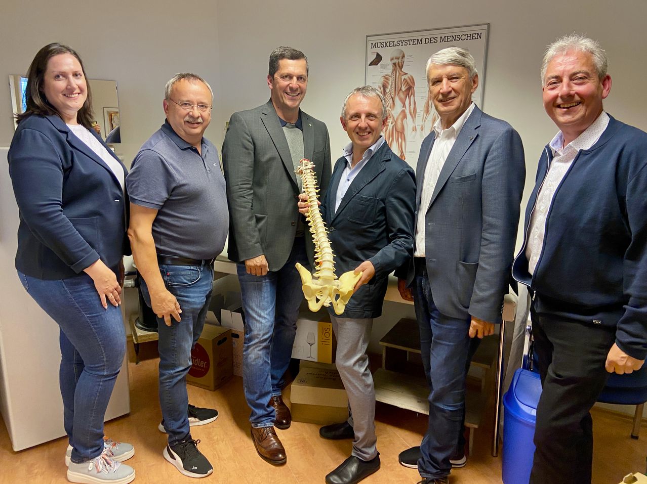 Sechs Männer in Anzügen posieren für ein Foto mit einem Modell einer menschlichen Wirbelsäule in einem Raum mit einer weißen Wand und einem Poster des menschlichen Muskelsystems.