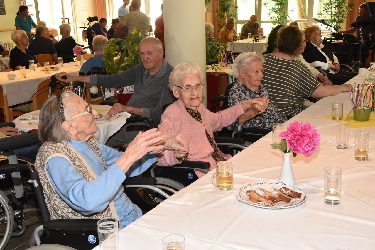 Eine Gruppe älterer Menschen in Rollstühlen klatscht in einem Essbereich. Es gibt einen Tisch mit einer Blumenvase, Brot und Getränken. Im Hintergrund sitzen weitere Menschen.
