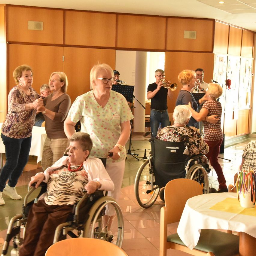 Eine Gruppe älterer Menschen, einige in Rollstühlen, tanzt und sozialisiert in einem Raum mit aufgebauten Musikinstrumenten im Hintergrund.