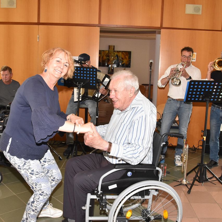 Ein älterer Mann im Rollstuhl lächelt und hält die Hand einer Frau. Hinter ihnen spielen Musiker Trompeten und Akkordeon. Ein Mikrofon auf einem Ständer steht vor ihnen.