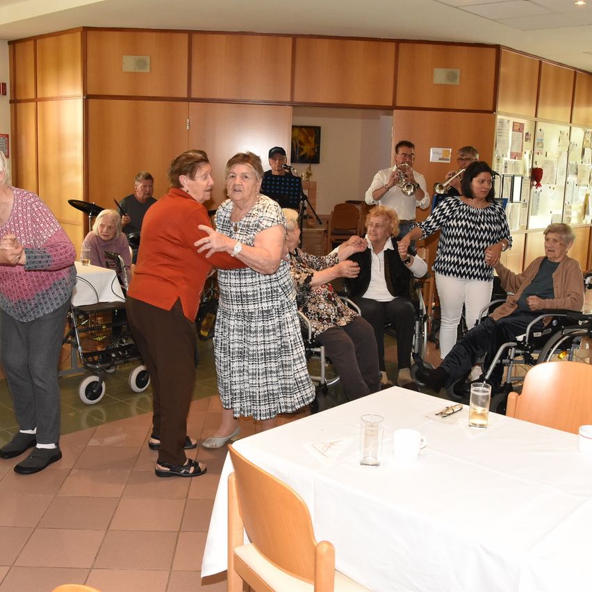 Mehrere ältere Frauen tanzen in einem Gemeinschaftsraum, einige sitzen in Rollstühlen. Einige Musiker spielen Instrumente und es gibt einen Tisch mit Getränken und einem Rollstuhl in der Nähe.