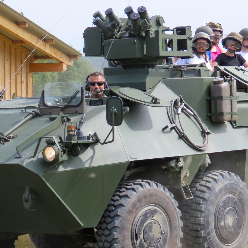 Ein grünes gepanzertes Fahrzeug mit montierten Waffen steht auf einem Grasfeld, auf dem mehrere Personen mit Helmen und Schutzbrillen sitzen.
