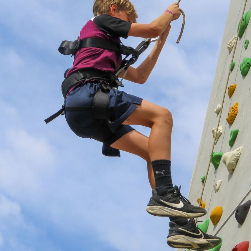 Ein junger Junge mit einem Gurt klettert an einer Kletterwand hoch. Er trägt Turnschuhe und Shorts und greift mit den Händen nach Halt.
