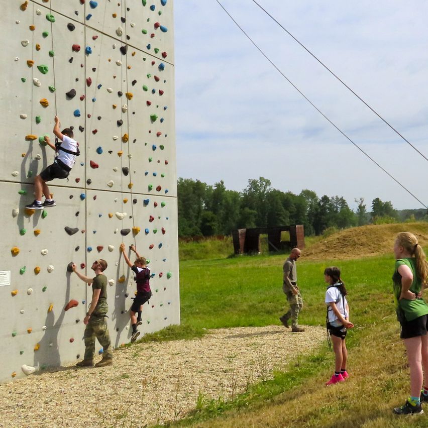 Eine Gruppe von Kindern und Erwachsenen klettert an einer Felswand im Freien. Die Wand ist hoch und hat viele farbige Griffe. Ein Mann geht in der Nähe vorbei. Im Hintergrund gibt es ein Grasfeld mit Bäumen und einem Gebäude.