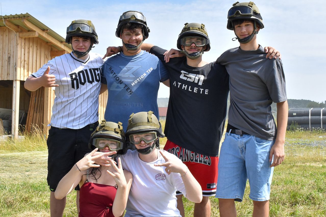 Fünf junge Personen stehen und sitzen auf einem Grasfeld und tragen Schutzausrüstung und Schutzbrillen. Sie posieren für ein Foto. Hinter ihnen steht ein Holzgebäude.