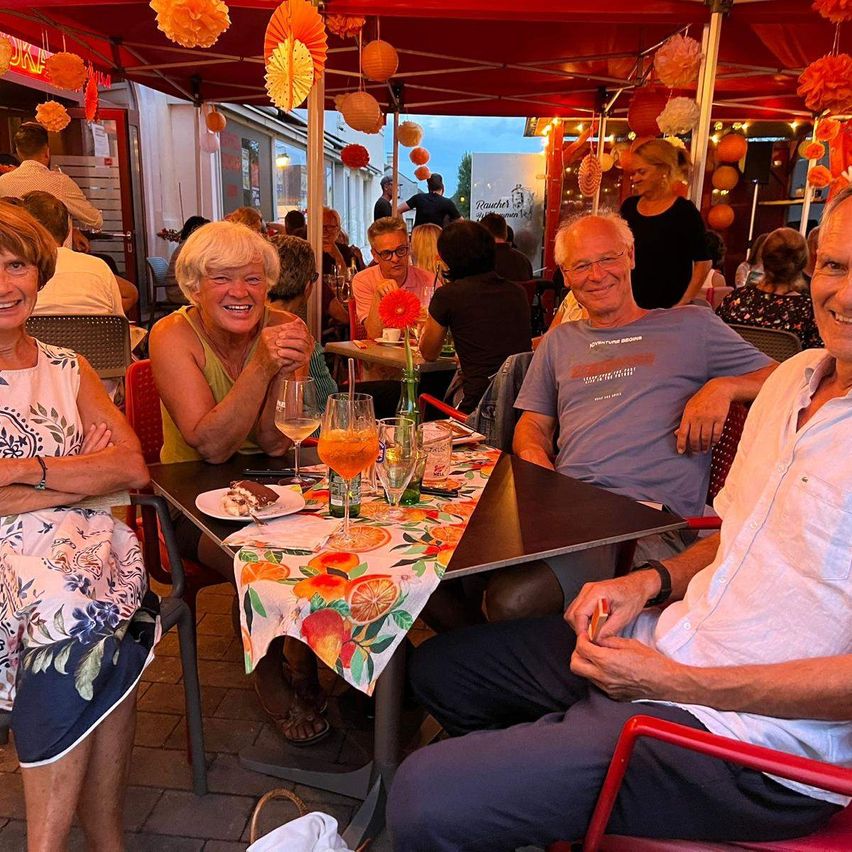 Eine Gruppe älterer Menschen lächelt und sitzt unter einem Zelt mit orangefarbenen Dekorationen. Sie essen mit Getränken und Essen auf dem Tisch. Im Hintergrund sitzen und stehen andere Menschen.