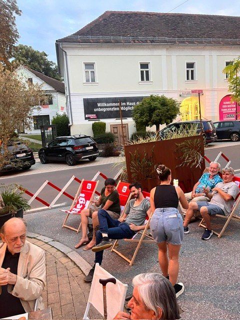 Eine Gruppe von Menschen sitzt auf Liegestühlen auf einer Straße. Es sind Autos geparkt und ein Gebäude mit einem Schild im Hintergrund.