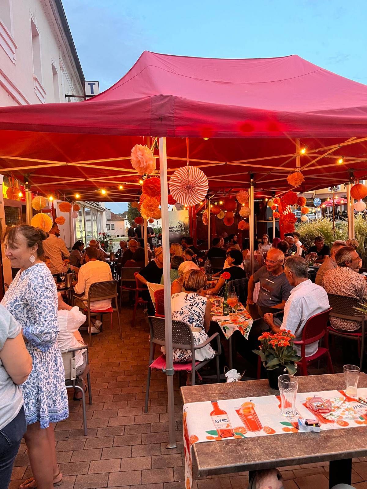 Ein Außenbereich mit rotem Zelt und Dekorationen. Die Leute sitzen an Tischen, einige mit Blumen und orangefarbenen Papierlaternen. Die Atmosphäre ist festlich mit warmer Beleuchtung.