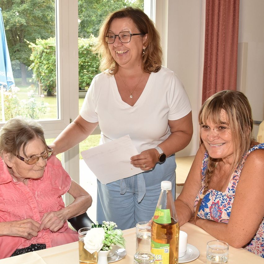 Drei Frauen sitzen an einem Tisch in einem Raum. Eine Frau hält ein Papier in der Hand und die anderen beiden lächeln. Sie haben Getränke und Blumen auf dem Tisch. Hinter ihnen befindet sich ein Fenster mit Vorhängen.