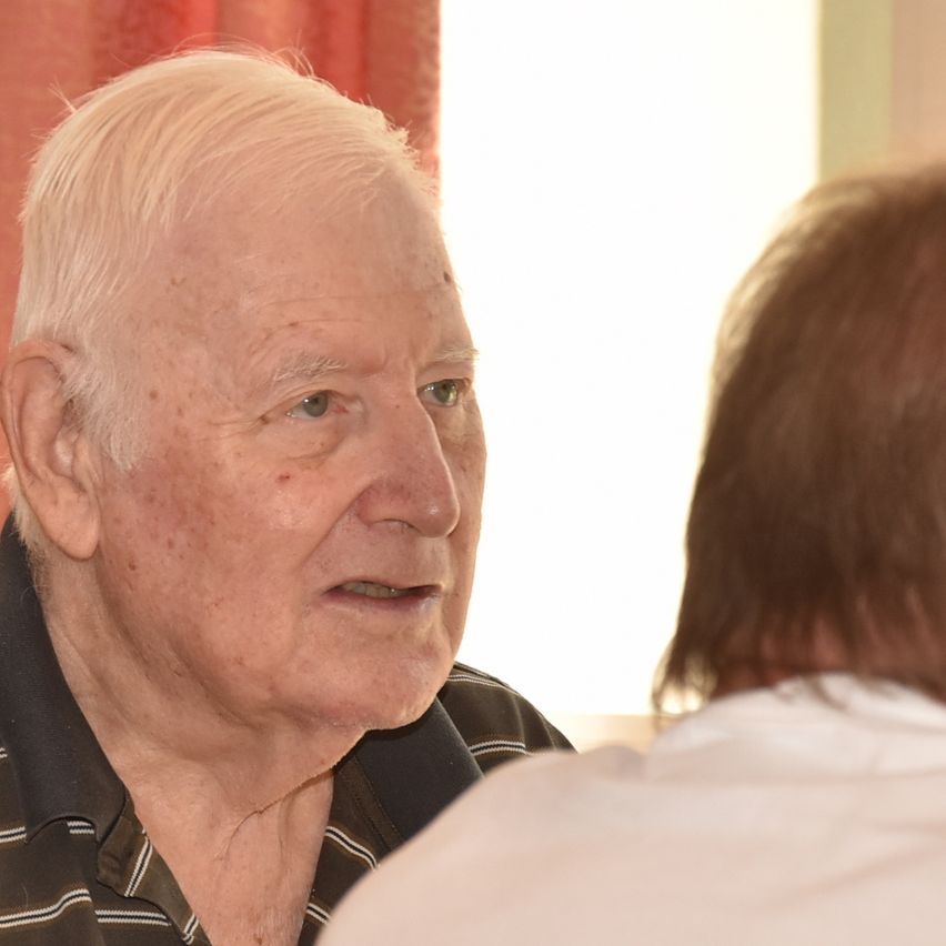 Ein älterer Mann mit weißen Haaren, Falten und einem schwarzen gestreiften Polo-Shirt spricht mit einem anderen Mann mit braunen Haaren, beide in einer Innenumgebung.