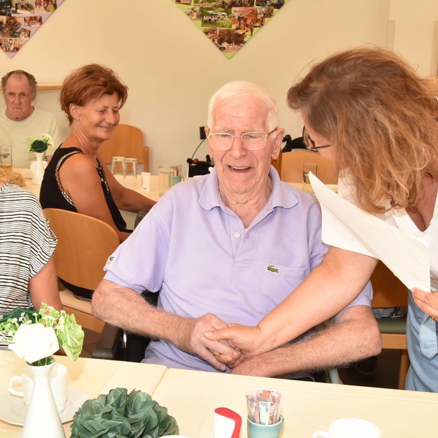 Ein älterer Mann im Rollstuhl, der ein lila Poloshirt trägt, hält die Hand einer Frau. Sie befinden sich in einem Essbereich, in dem mehrere Menschen an Tischen sitzen.