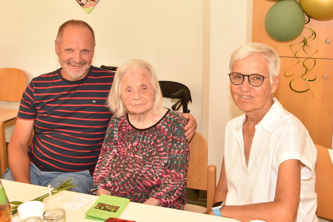 Drei Menschen sitzen lächelnd am Tisch. Eine ältere Frau in der Mitte mit einem Blumenhemd, ein Mann mit einem gestreiften Shirt und ein weiterer Mann in einem weißen Hemd. Hinter ihnen eine Wand mit Luftballons und ein Stuhl.