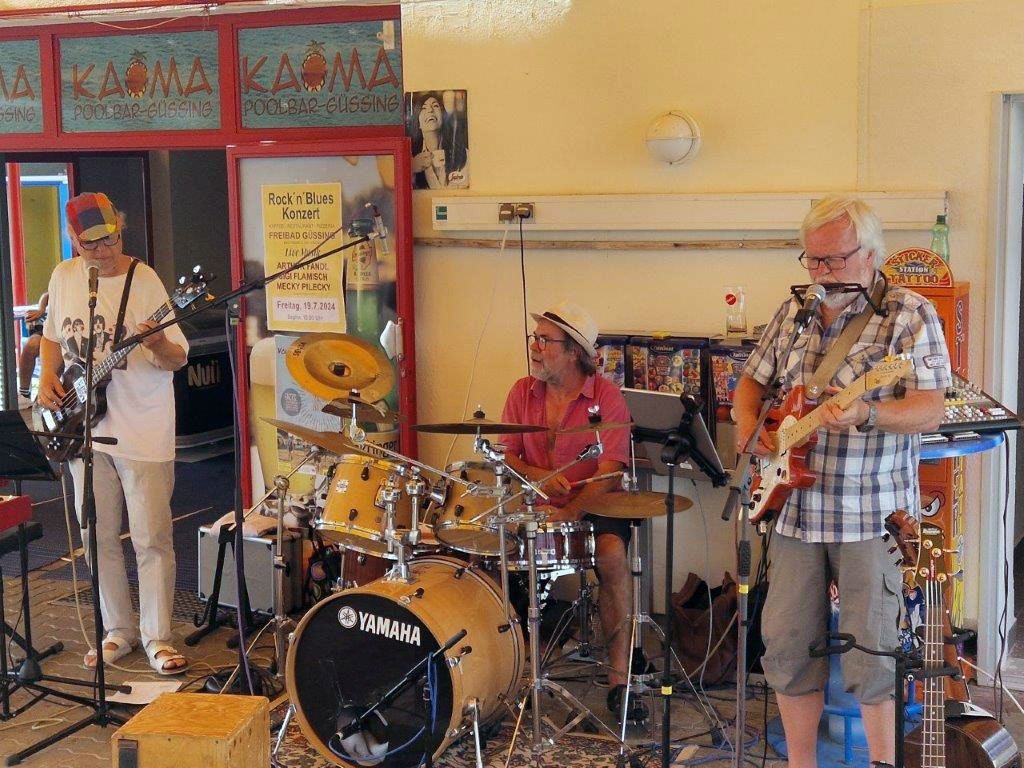 Eine Gruppe Musiker tritt im Freien auf. Einer spielt Gitarre und singt, ein anderer Schlagzeug und ein Dritter Bassgitarre. Das Schild im Hintergrund lautet 'Rock n Blues Konzert'.