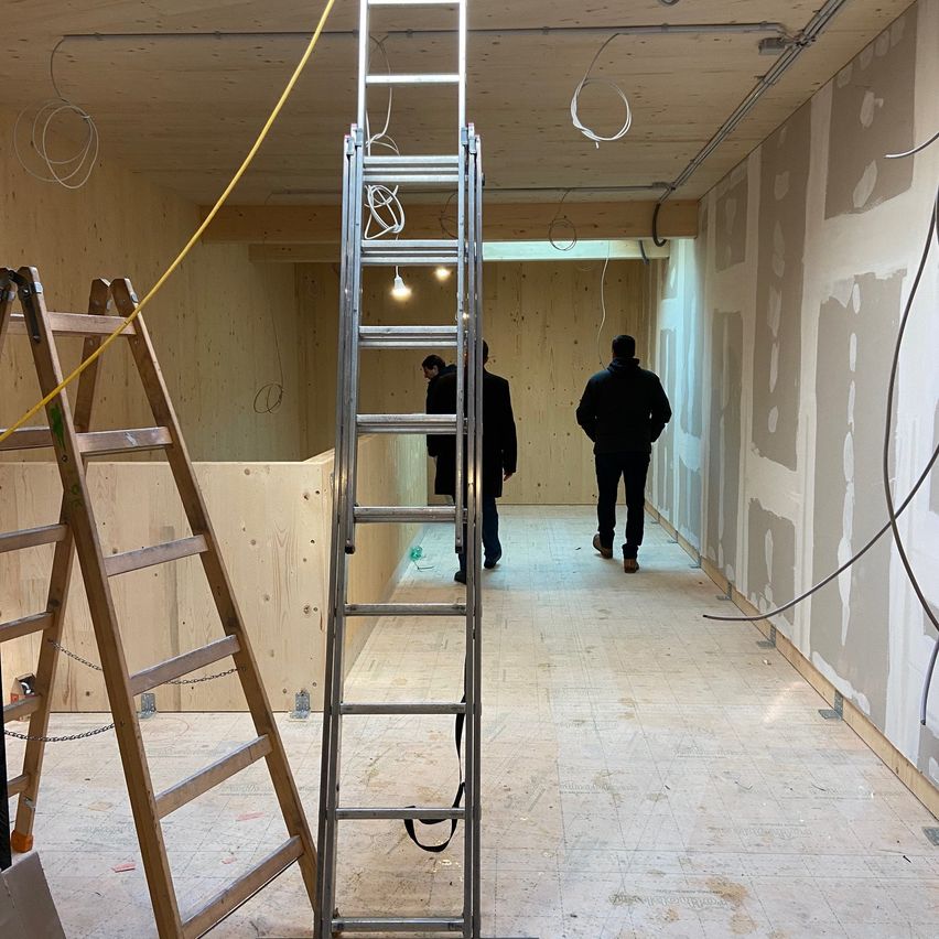 Ein Innenraum im Bau mit einer hölzernen Wand und unfertigem Boden, zwei Personen gehen einen Korridor entlang. Es gibt eine Leiter, Kabel und eine hölzerne Leiter auf der linken Seite.