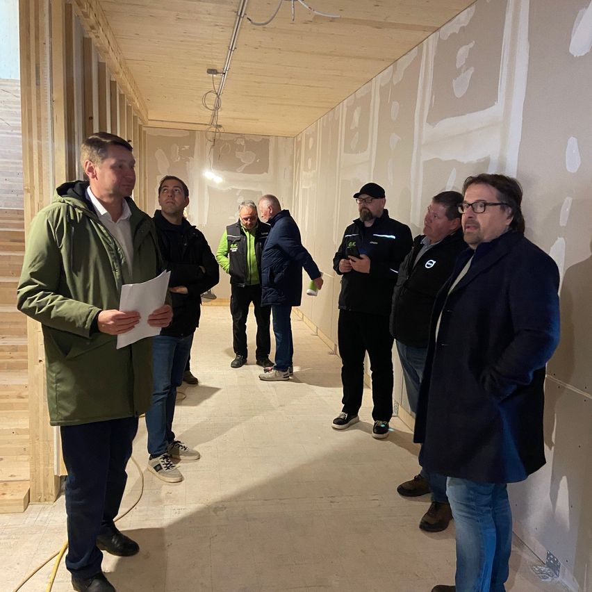 Eine Gruppe von Männern, die Winterkleidung tragen, steht in einem Korridor, wobei einer einen Zettel in der Hand hält. Sie diskutieren wahrscheinlich etwas.