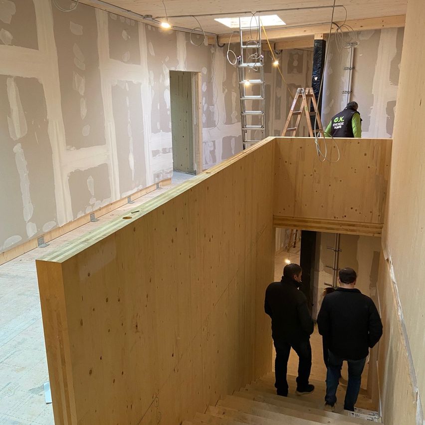 Drei Männer stehen auf einer hölzernen Treppe. Ein Mann steht auf der obersten Stufe, ein anderer darunter. Der dritte Mann steht im unteren Stockwerk. Der Raum ist im Bau.