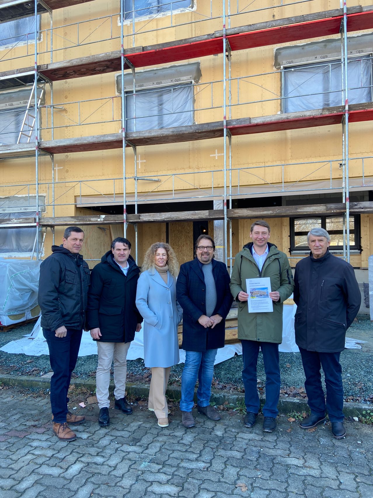Sechs Menschen stehen vor einem im Bau befindlichen Gebäude. Ein Mann hält ein Zertifikat. Sie sind alle warm gekleidet.