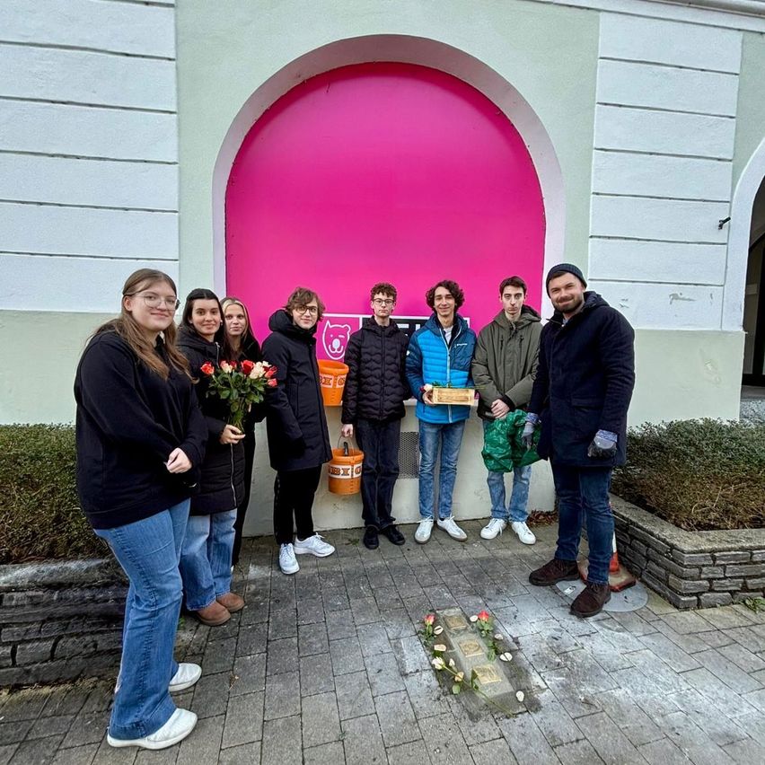 Eine Gruppe von Menschen in Winterkleidung steht vor einem rosafarbenen Torbogen, einige halten Blumen und Eimer. Sie lächeln und scheinen für ein Foto zu posieren.