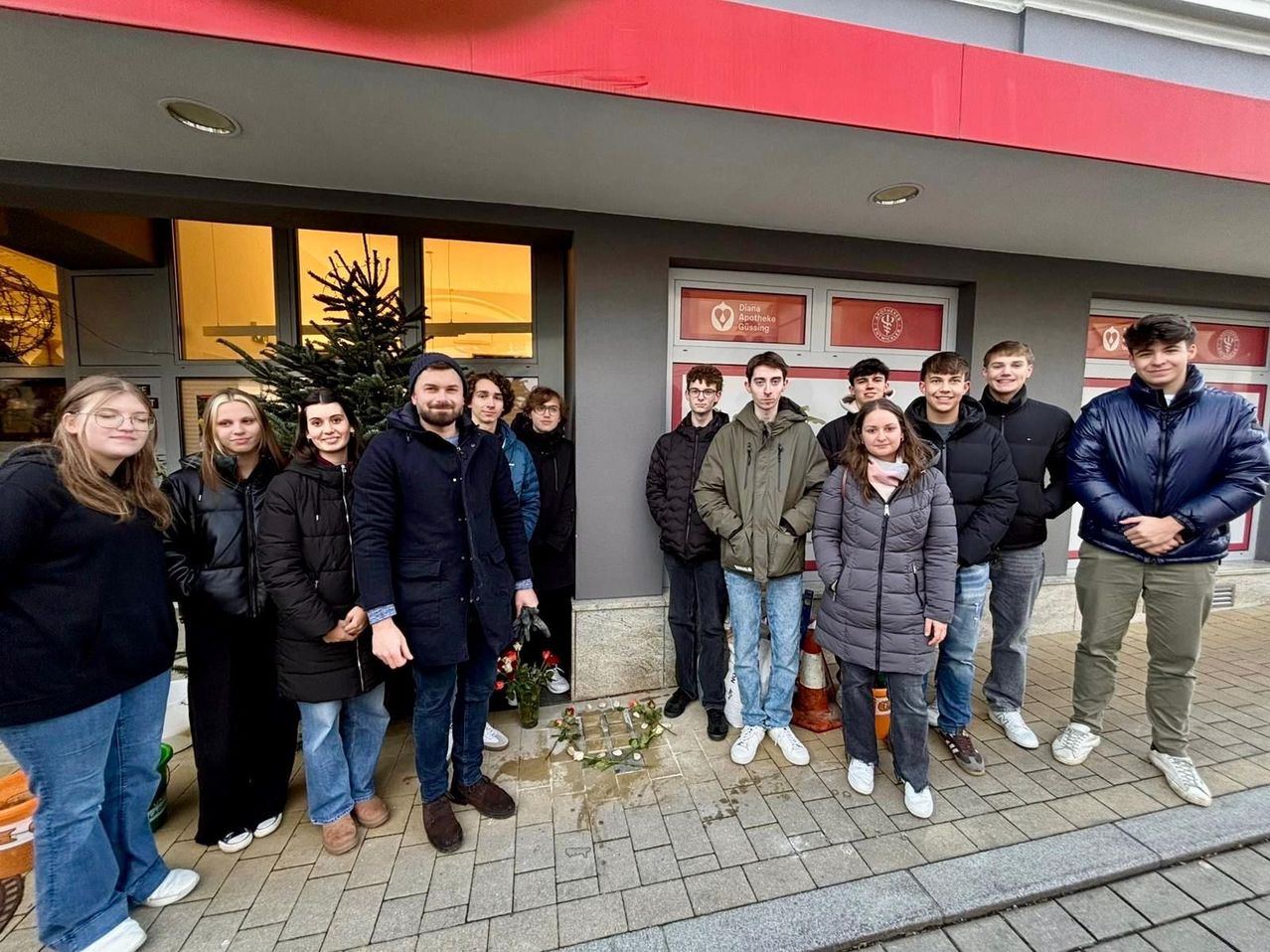Eine Gruppe junger Menschen posiert für ein Foto vor einem Gebäude, vor dem ein Weihnachtsbaum und Blumen aufgestellt sind.