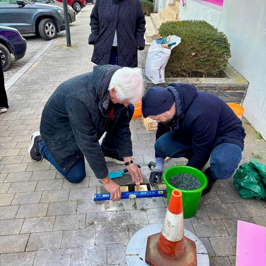 Zwei Männer knien auf einem gepflasterten Bürgersteig, vielleicht um einen Bordsteinmarker zu installieren. Einer hält ein Maßband. Ein Verkehrskegel und ein grüner Eimer sind in der Nähe. Hinter ihnen befindet sich ein Gebäude mit Treppen. Autos sind auf der Seite geparkt.