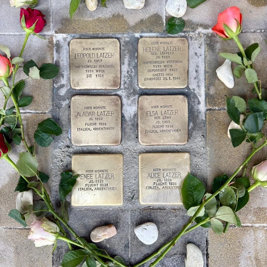 Sechs Gedenksteine auf einem Ziegelboden, jeder mit einem Namen und Daten graviert. Umgeben von roten und rosa Rosen und kleinen Steinen. Die Steine tragen Namen wie Helene Latzer, Leopold Latzer und andere, zusammen mit Geburts- und Sterbejahren.
