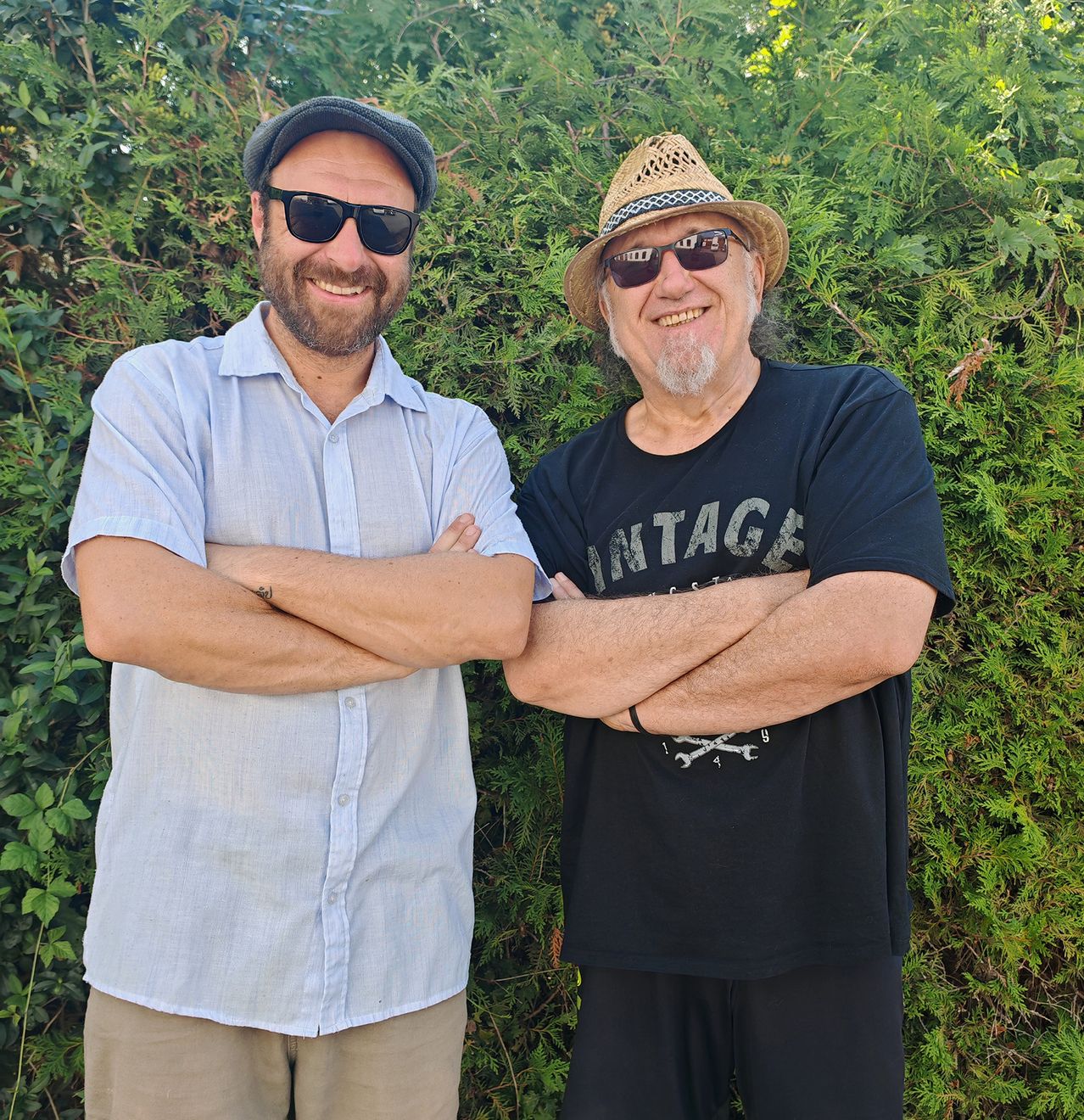 Zwei Männer stehen nebeneinander, lächeln und posieren für ein Foto. Der Mann auf der linken Seite trägt einen Hut und ein hellblaues Hemd, während der Mann auf der rechten Seite einen Strohhut und ein schwarzes T-Shirt trägt. Sie haben beide die Arme verschränkt.