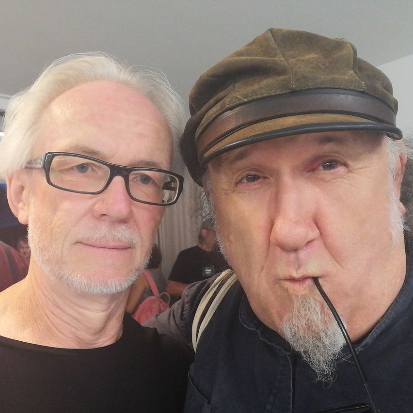 Zwei Männer stehen eng beieinander. Der Mann auf der linken Seite trägt eine Brille und ein schwarzes T-Shirt. Der Mann auf der rechten Seite trägt einen Hut und ein schwarzes T-Shirt mit einer Zigarette im Mund. Hinter ihnen stehen Menschen und eine Frau trägt eine rosa Tasche.