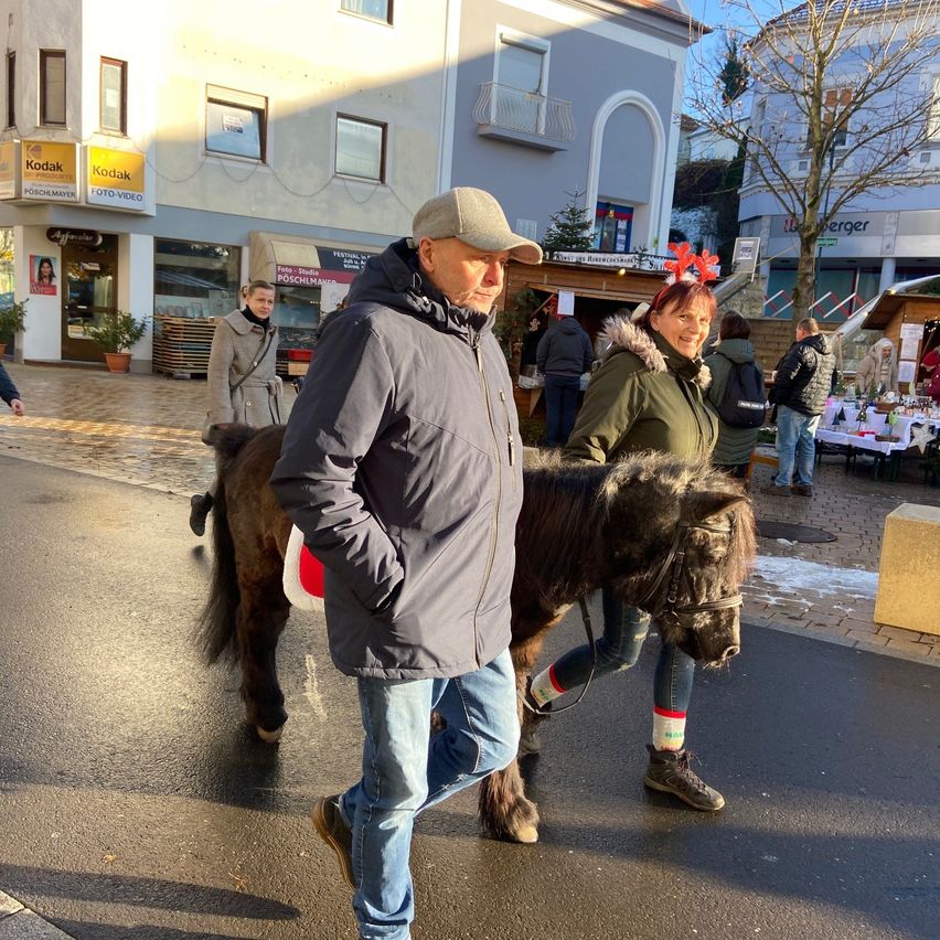 Ein Mann und eine Frau führen ein kleines Pferd die Straße entlang. Hinter ihnen befinden sich Gebäude mit Kodak-Schildern und einem Weihnachtsmarkt.
