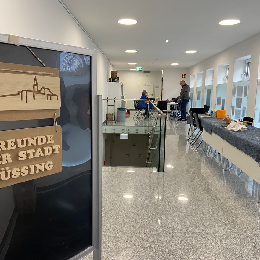 Ein Flur in einem Gebäude mit sitzenden Personen an Tischen und stehenden Personen. Ein Schild steht 'runde stadt bussing'.