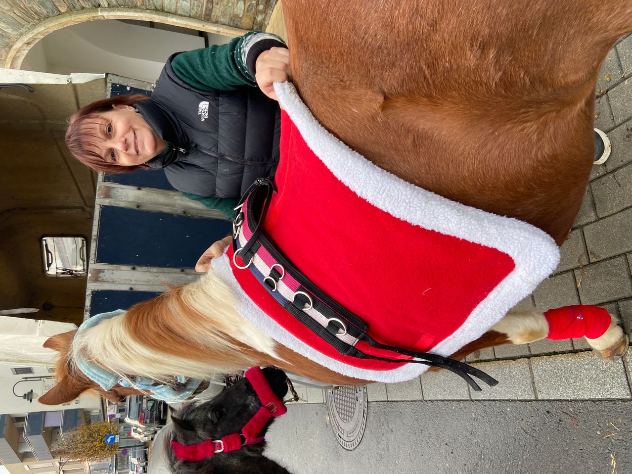 Eine Frau mit kurzen roten Haaren lächelt, während sie die Zügel eines in einem roten und weißen Mantel gekleideten Pferdes hält, das auf einem Bürgersteig steht.