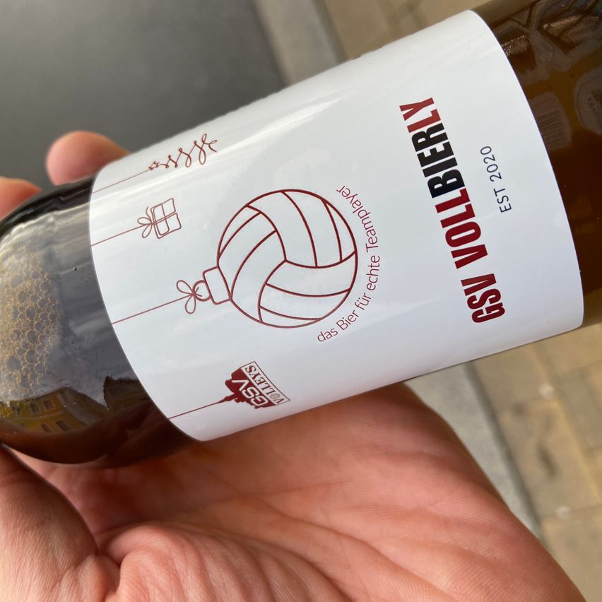 Eine Nahaufnahme einer Hand, die eine Flasche GSI Vollbierly Bier aus dem Jahr 2020 hält, mit einem roten Volleyball und Geschenken.