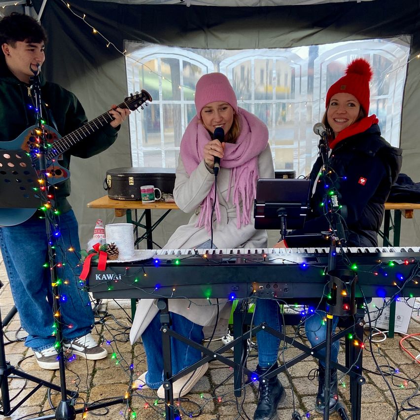 Drei Personen treten auf der Bühne auf, geschmückt mit Weihnachtslichtern. Ein Mann spielt Gitarre, während zwei Frauen singen, eine mit rosa Mütze und Schal, die andere mit roter Mütze.