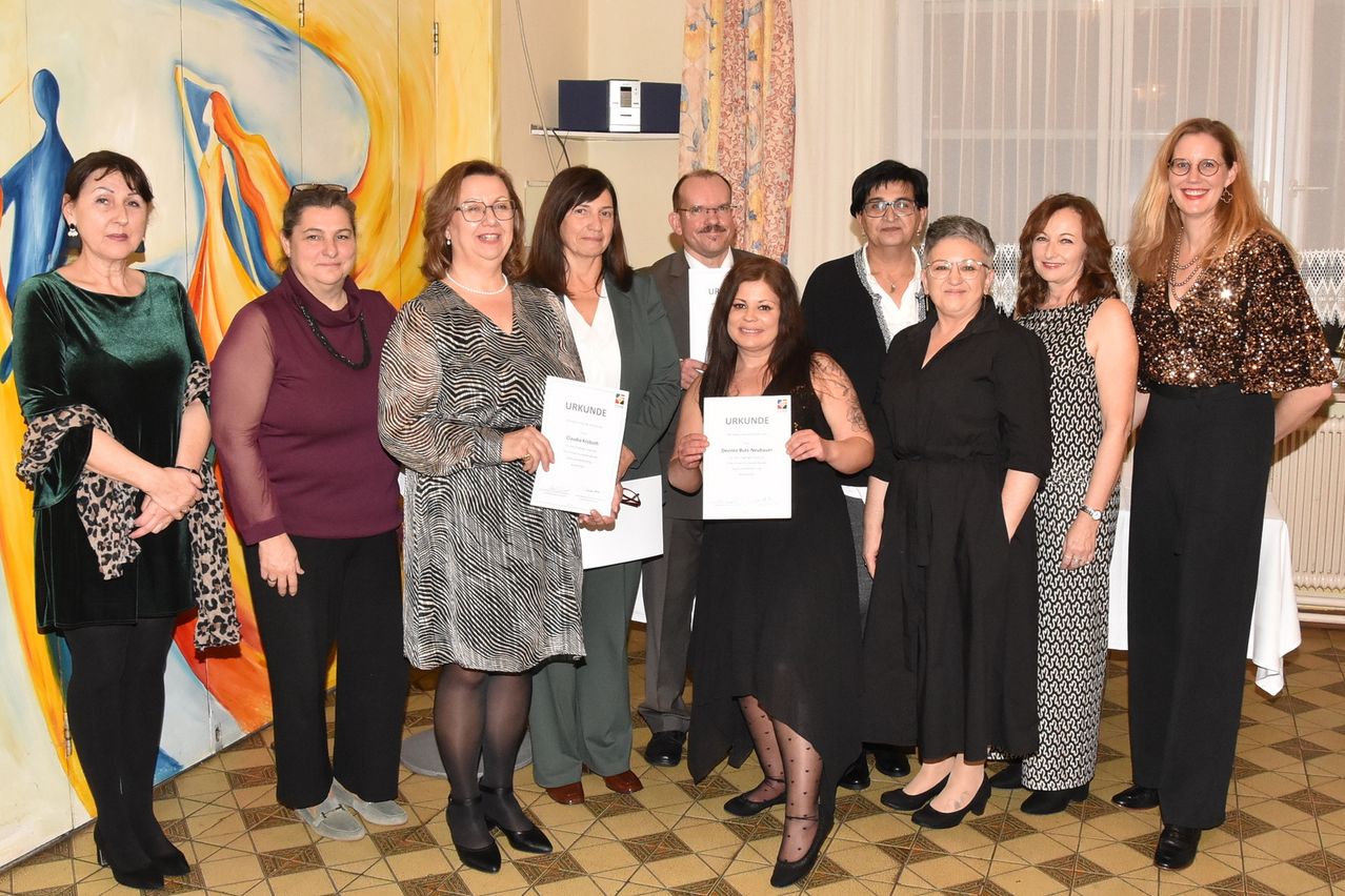 Eine Gruppe von Frauen und Männern steht in einem Raum und hält Urkunden in den Händen. Zwei Frauen in der Mitte halten Urkunden mit dem Wort 'Urkunde' darauf. Der Raum hat weiße Vorhänge und eine dekorative Malerei an der Wand.