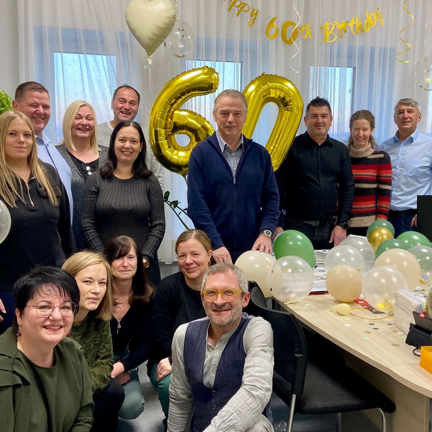 Eine Gruppe von Menschen versammelt sich drinnen zu einer Geburtstagsfeier, mit Luftballons und einer Nummer 60 Dekoration im Hintergrund.