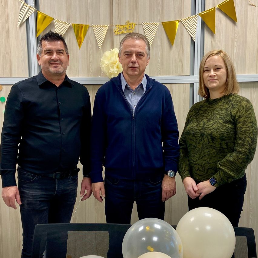 Drei Menschen stehen vor einer Wand mit einer Geburtstagsdekoration. Der Mann auf der linken Seite trägt ein schwarzes Hemd, der Mann in der Mitte trägt eine blaue Jacke und die Frau auf der rechten Seite trägt einen grünen Pullover. Sie stehen neben einem Tisch mit drei weißen Luftballons.