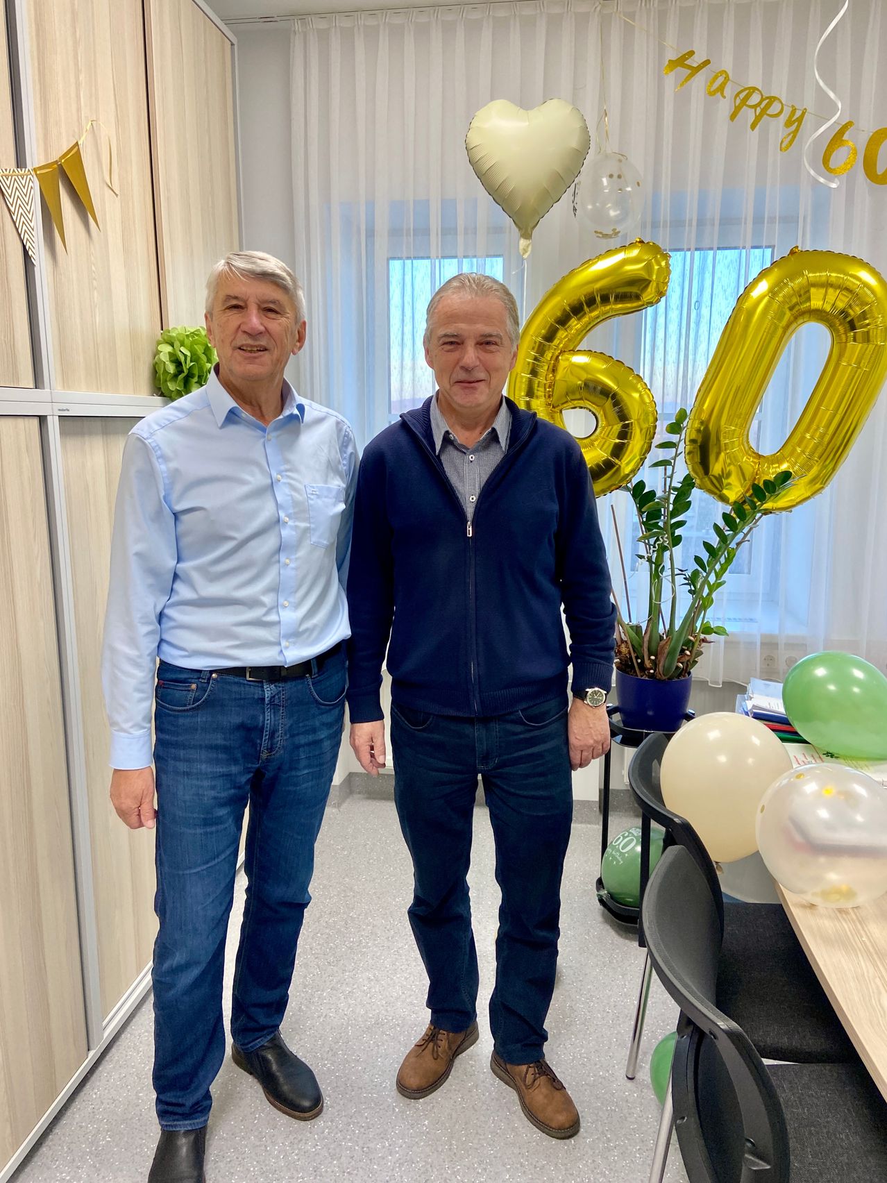 Zwei Männer stehen in einem Büro und lächeln für ein Foto. Einer trägt ein hellblaues Hemd und der andere eine dunkelblaue Jacke. Hinter ihnen befinden sich Luftballons mit der Nummer 60 und eine Topfpflanze.